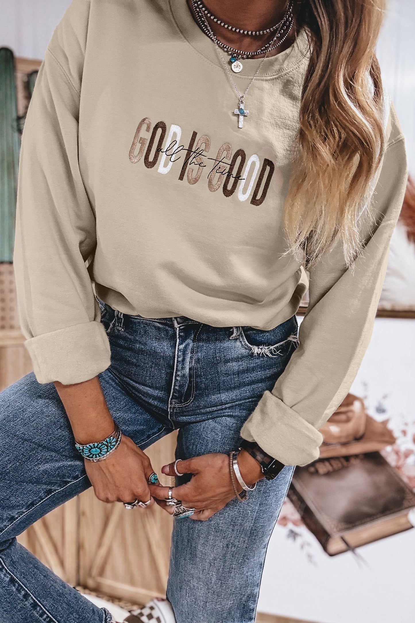 Close-up of raised embroidered God Is Good All The Time lettering on sand crewneck sweatshirt styled with jeans