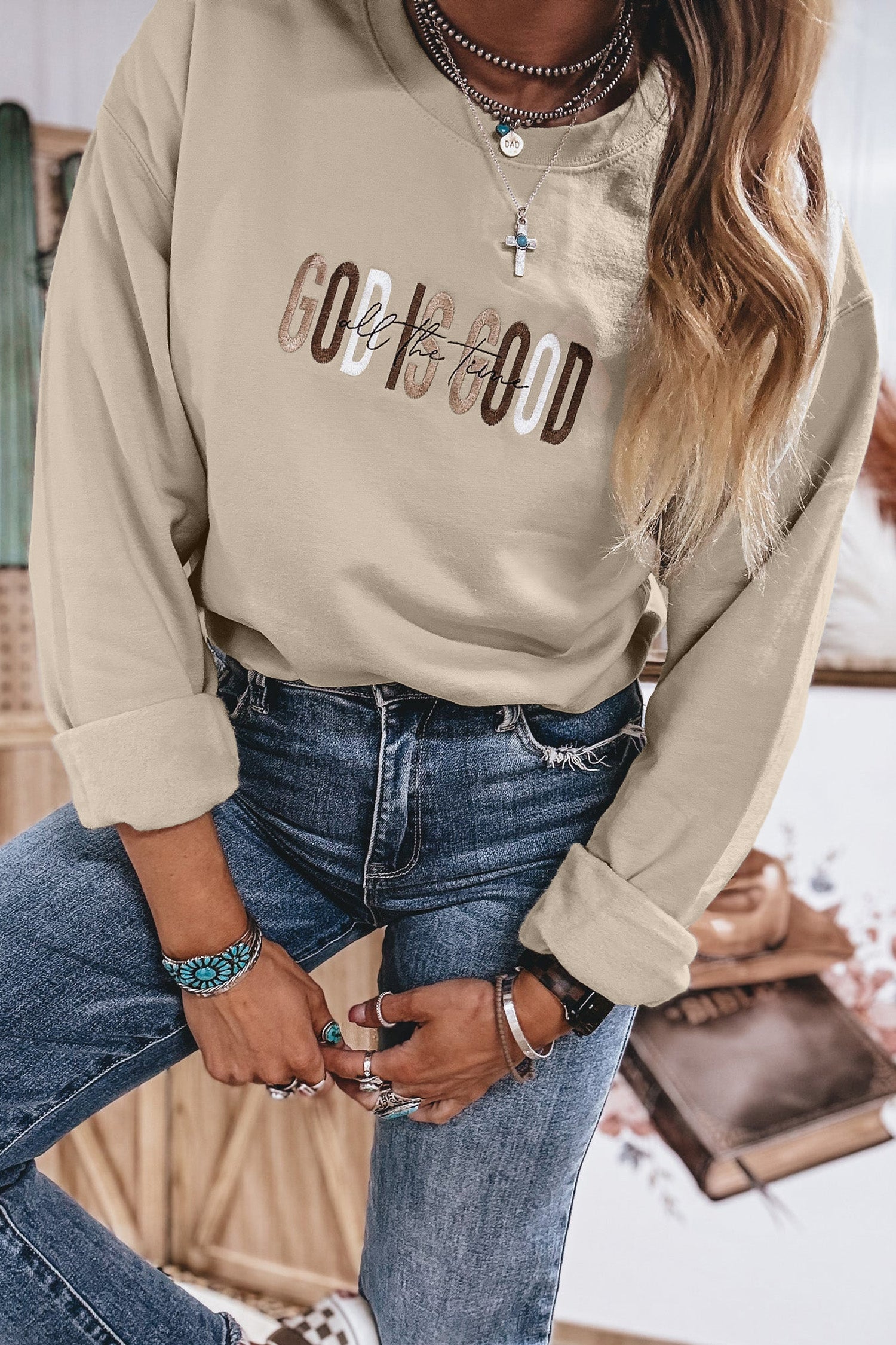 Close-up of raised embroidered God Is Good All The Time lettering on sand crewneck sweatshirt styled with jeans